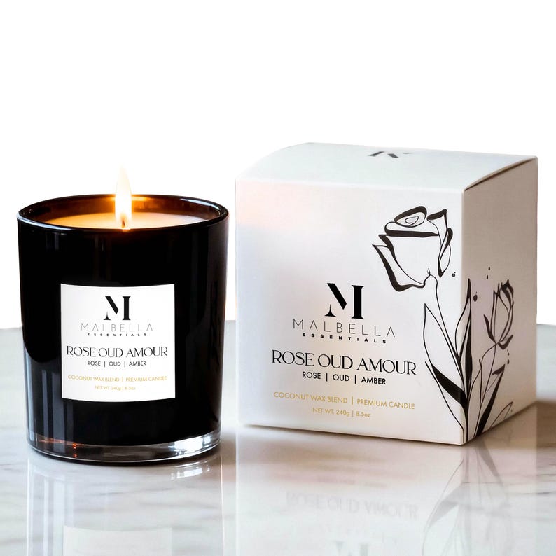 May include: A lit black glass candle with a white label that reads "MALBELLA ESSENTIALS ROSE OUD AMOUR ROSE | OUD | AMBER". A white box with the same text and a rose illustration is next to the candle. The candle is 3.5 oz.