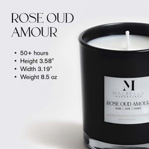 May include: A black glass candle with a white label that reads "ROSE OUD AMOUR." The candle has a white wax interior and a wick. The label also includes the brand name "MALBELLA ESSENTIALS." The candle is 3.58 inches tall and 3.19 inches wide.