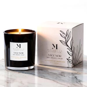 May include: A lit black glass candle with the text "SAGE NOIR" and a white label. The candle is next to a white box with the same text and a botanical illustration. The candle is a coconut wax blend and weighs 140g / 5 oz.