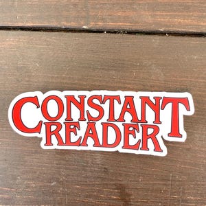 May include: A white sticker with the words "CONSTANT READER" in red, bold, serif font. The sticker has a red border and is set against a dark wood grain background. The text is in two lines.