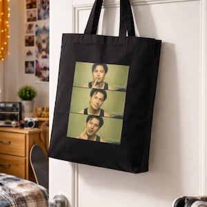 Può includere: Borsa tote nera con un'immagine rettangolare di una persona in tre pannelli. La persona indossa una canotta nera e fa un gesto con il dito sulle labbra. La borsa è appesa a una porta bianca.