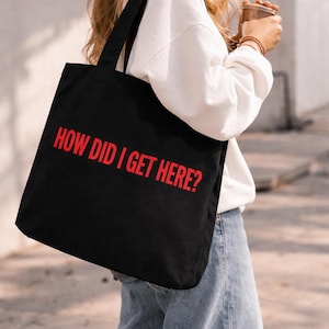 May include: Black tote bag with the red text "HOW DID I GET HERE?". The bag has black handles and is being carried over the shoulder. The person is wearing a white sweatshirt and blue jeans.