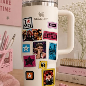 Puede incluir: Un vaso Stanley de color crema con asa y pajita transparente, decorado con varios adhesivos rectangulares y cuadrados que muestran imágenes de bandas y textos como 5SOS, IS NOT OK, BITE THE APPLE BABY e IRWIN 94 sobre el cuerpo del vaso.