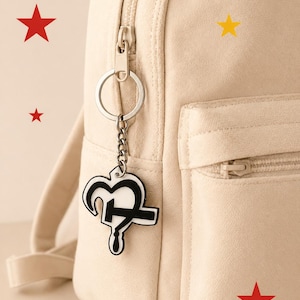 Può includere: Un portachiavi a forma di cuore bianco e nero con un design a punto interrogativo è appeso a una catena d'argento attaccata a uno zaino color crema. Stelle rosse e gialle sono sparse intorno allo zaino.