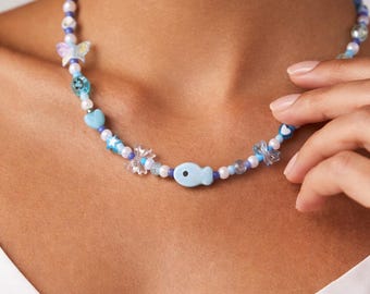 Op de afbeelding: Een kralenketting met een lichtblauwe vishanger, vlinderbedels en hartvormige kralen. De ketting heeft een mix van blauwe, witte en transparante kralen. De ketting wordt om de nek gedragen.