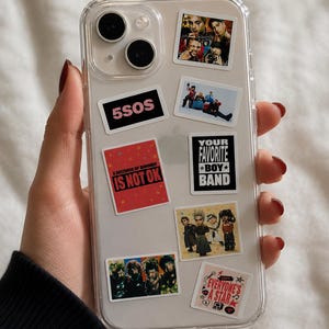 Può includere: Custodia trasparente per telefono decorata con adesivi con foto di band e testo. Gli adesivi includono "5SOS", "6 SECONDS OF SUMMER IS NOT OK", "YOUR FAVORITE BOY BAND" e "EVERYONE'S A STAR". La custodia è tenuta da una persona.