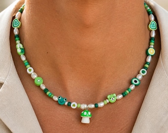 May include: A beaded necklace featuring green and white beads, with charms including a mushroom, frog, bear, and fruit slices. The necklace is composed of white pearls and green beads, creating a playful and colorful accessory.