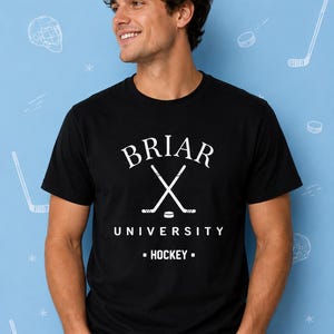 May include: A black short-sleeved t-shirt featuring white text that reads BRIAR UNIVERSITY HOCKEY with a graphic of two crossed hockey sticks and a puck in the centre. The shirt has a crew neckline and is made of a solid black fabric.