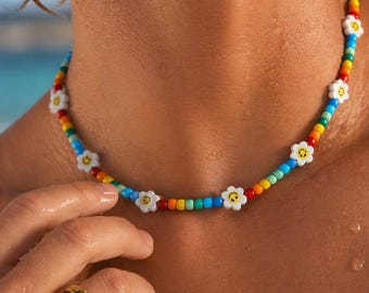 May include: A colorful beaded necklace with a rainbow pattern and white daisy flower accents. The necklace is worn around the neck. A gold ring is on the finger.