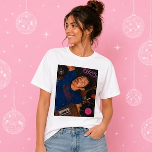 Può includere: T-shirt bianca con grafica colorata con la scritta "DISCO" e l'immagine di un uomo. La maglietta è indossata da una persona con uno chignon, su sfondo rosa decorato con palle da discoteca.