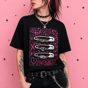 May include: Black t-shirt with a pink graphic featuring three safety pins and the text "YUNGBLUD". The design includes hearts, skulls, and crosses. The model is wearing a black choker and a silver chain.