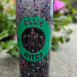 May include: A clear tumbler filled with black glitter and colorful specks. A green circle with the words "BASIC WITCH" surrounds a witch silhouette. The tumbler is a fun, festive item.