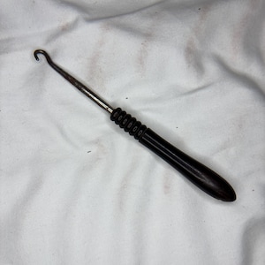 May include: A dark wooden crochet hook with a metal shaft and a curved hook end. The handle is a dark brown color and has a series of ridges near the metal shaft. The hook is designed for pulling loops of yarn through other loops.