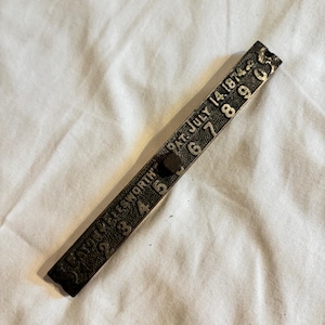 May include: A vintage metal ruler with embossed numbers and text, including "PAT. JULY 14, 1874" and "AYON KELLSWORTH". The ruler is dark brown and lies on a white fabric surface.