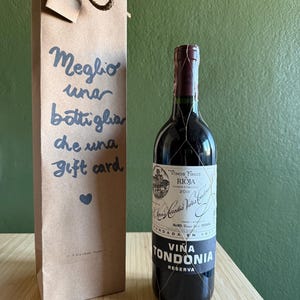 以下が含まれることがあります： Viña Tondonia Reserva Riojaワインのボトルと茶色の紙袋。ワインボトルはワイヤーメッシュで包まれ、ラベルが付いています。ギフトバッグには「Meglio una bottiglia che una gift card」と書かれています。