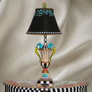 May include: A decorative table lamp featuring a black lampshade with teal scrollwork and a checkered trim. The base is a multi-colored urn shape with gold, white, and black patterns, accented by bright teal handles and a square base with a checkered border.