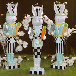 May include: Three ceramic bunny figurines dressed as Easter nutcrackers. Each bunny is wearing a blue and white checkered suit, a black top hat, and holding a carrot. The bunnies are standing on a black and white checkered base.