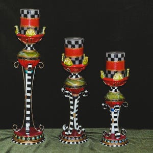 May include: Three decorative candle holders with a whimsical design. Each holder features a red and black checkered candle cup, a green and gold midsection, and a black and white striped base. The tallest holder is approximately 18 inches tall.