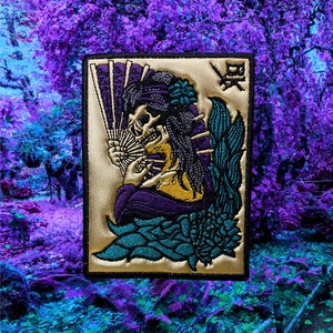 May include: A gold embroidered patch depicting a skull holding a fan, dressed in a purple and black kimono, sitting on a blue and green flower. The patch has a gold border and the Japanese character "鬼" in the top right corner.