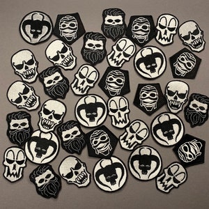 May include: A collection of black and white iron-on patches featuring various skull designs. The patches are arranged in a random pattern on a grey background.