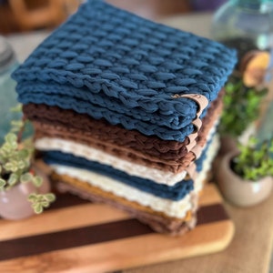 May include: A stack of crocheted pot holders in various colors, including blue, brown, white, and yellow. The pot holders are stacked on a wooden cutting board.