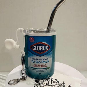 May include: A clear plastic container with a Clorox Disinfecting Wipes To Go Pack label. The container has a metal straw and a metal keychain attached. The container is sitting on a white surface with a sunflower design.