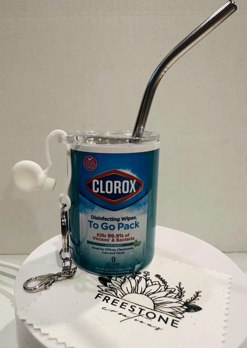 May include: A clear plastic container with a Clorox Disinfecting Wipes To Go Pack label. The container has a metal straw and a metal keychain attached. The container is sitting on a white surface with a sunflower design.