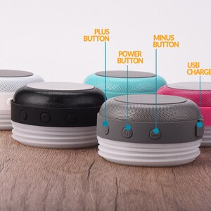 May include: Four portable Bluetooth speakers in different colors: white, black, gray, and pink. Each speaker has a USB charging port, power button, plus and minus buttons, and a textured top.