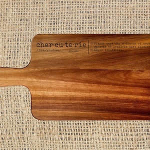 May include: Wooden charcuterie board with a handle and a hole for hanging. The board features the word "charcuterie" with its pronunciation and definition. The board is made of a rich, warm-toned wood.
