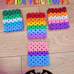 May include: Four colorful beaded cuffs arranged on a wooden surface. The cuffs display the colors of various pride flags, including rainbow, lesbian, and bisexual flags. The text "PRIDE FLAG CUFFS" is at the top.