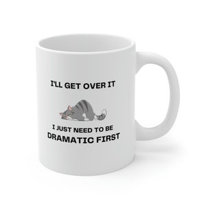 May include: White ceramic coffee mug with a black and white illustration of a cat. The text on the mug reads "I'll get over it I just need to be dramatic first."