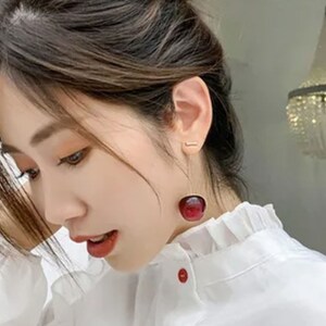 May include: A woman wearing a white collared shirt with a ruffled neckline. She is wearing a pair of red cherry earrings.