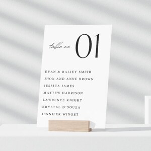 May include: A white table number card with the number "01" in a bold, black font. The words "table no." are written in a cursive font. Below the number are names, including "EVAN & BALIEY SMITH" and "JHON AND ANNE BROWN". The card is held by a light wood block.