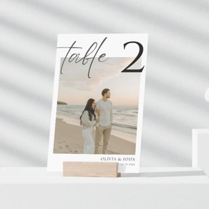 以下が含まれることがあります： 白いテーブルナンバーカードに、黒い書体で「テーブル2」と書かれています。カードには、ビーチにいるカップルの写真と、下部に「Olivia & John 10.14.2024」というテキストが書かれています。