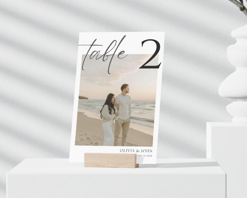 May include: White table number card with black calligraphy that says "Table 2". The card has a photo of a couple on a beach with the text "Olivia & John 10.14.2024" at the bottom.