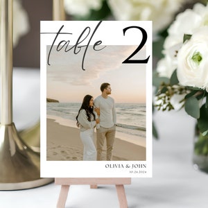 May include: A white table number card with black calligraphy that reads "table 2". The card has a photo of a couple on a beach at sunset. The text below the photo reads "Olivia & John 10.24.2024".