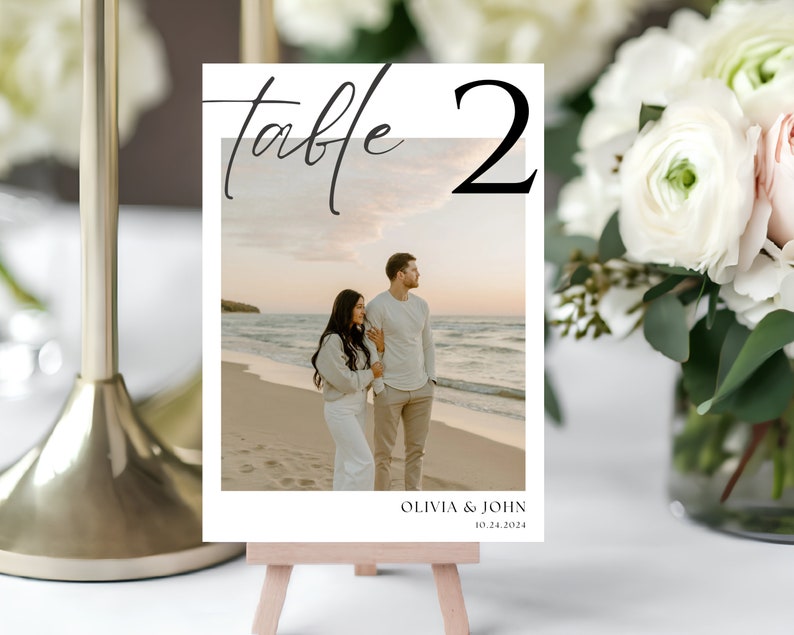 May include: A white table number card with black calligraphy that reads "table 2". The card has a photo of a couple on a beach at sunset. The text below the photo reads "Olivia & John 10.24.2024".