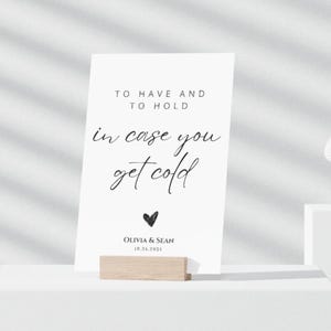 May include: White card with black text that reads "To have and to hold in case you get cold" with a small black heart and the text "Olivia & Sean 10.24.2023" below. The card is sitting on a wooden stand.
