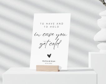 May include: White card with black text that reads "To have and to hold in case you get cold" with a small black heart and the text "Olivia & Sean 10.24.2023" below. The card is sitting on a wooden stand.