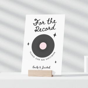 Puede incluir: Un letrero blanco con el texto "For the Record" y un gráfico de disco de vinilo. El letrero dice "Please Sign Our Guestbook" e incluye los nombres "Emily & Jackob". El letrero está sujeto por un soporte de madera.