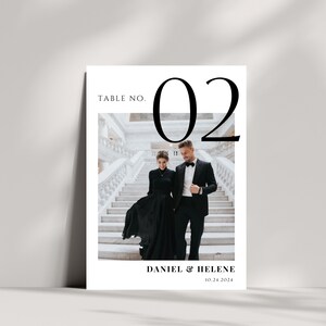May include: A wedding table number card with the number 02 in large black font. The card features a photo of a couple in black formal attire walking down a white staircase. The text "Daniel & Helene 10.24.2024" is printed below the photo.