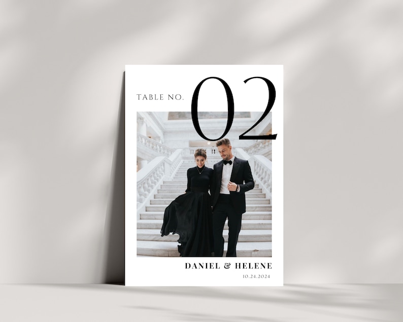 May include: A wedding table number card with the number 02 in large black font. The card features a photo of a couple in black formal attire walking down a white staircase. The text "Daniel & Helene 10.24.2024" is printed below the photo.