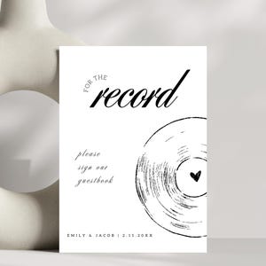 May include: A white sign with black text that reads "For the record, please sign our guestbook."  A black and white illustration of a vinyl record with a small heart in the centre is below the text. The text "Emily & Jacob | 2.15.20XX" is at the bottom of the sign.
