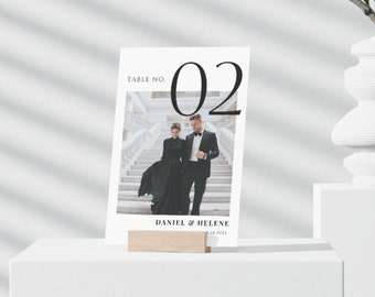 May include: A white card with black text that reads "Table No. 02" and a photo of a couple in black formal wear standing on a staircase. The text "Daniel & Helene" and the date "12.24.2021" are also on the card.
