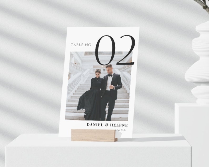 May include: A white card with black text that reads "Table No. 02" and a photo of a couple in black formal wear standing on a staircase. The text "Daniel & Helene" and the date "12.24.2021" are also on the card.