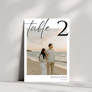 May include: A white table number card with the text "Table 2" in black. A photo of a couple walking on a beach at sunset is in the background. The text "Olivia & John 10.24.2024" is at the bottom of the card.