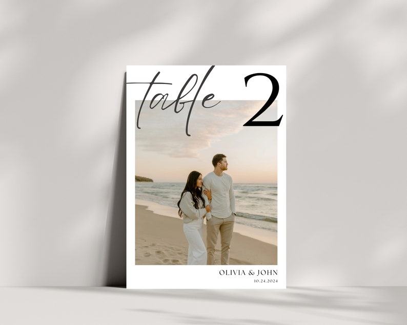 May include: A white table number card with the text "Table 2" in black. A photo of a couple walking on a beach at sunset is in the background. The text "Olivia & John 10.24.2024" is at the bottom of the card.
