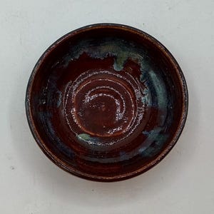 May include: A handmade ceramic bowl with a dark brown interior and a dark blue and green rim. The bowl has a glossy finish and a circular design in the center. The bowl is perfect for serving food.
