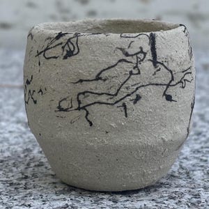 May include: A small, handmade ceramic cup with a rough, textured surface. The cup is a light beige color and is decorated with black, abstract line drawings. The cup has a rounded shape and a slightly flared rim, suitable for holding beverages.