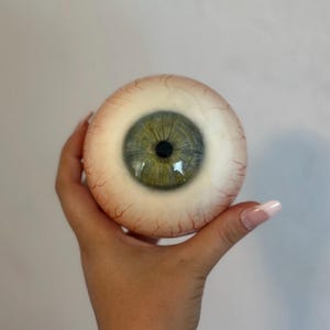 Puede incluir: Un ojo protésico realista y de gran tamaño con un iris verde y una pupila negra. La esclerótica es blanquecina con venas rojas. El ojo se sostiene en una mano sobre un fondo liso.